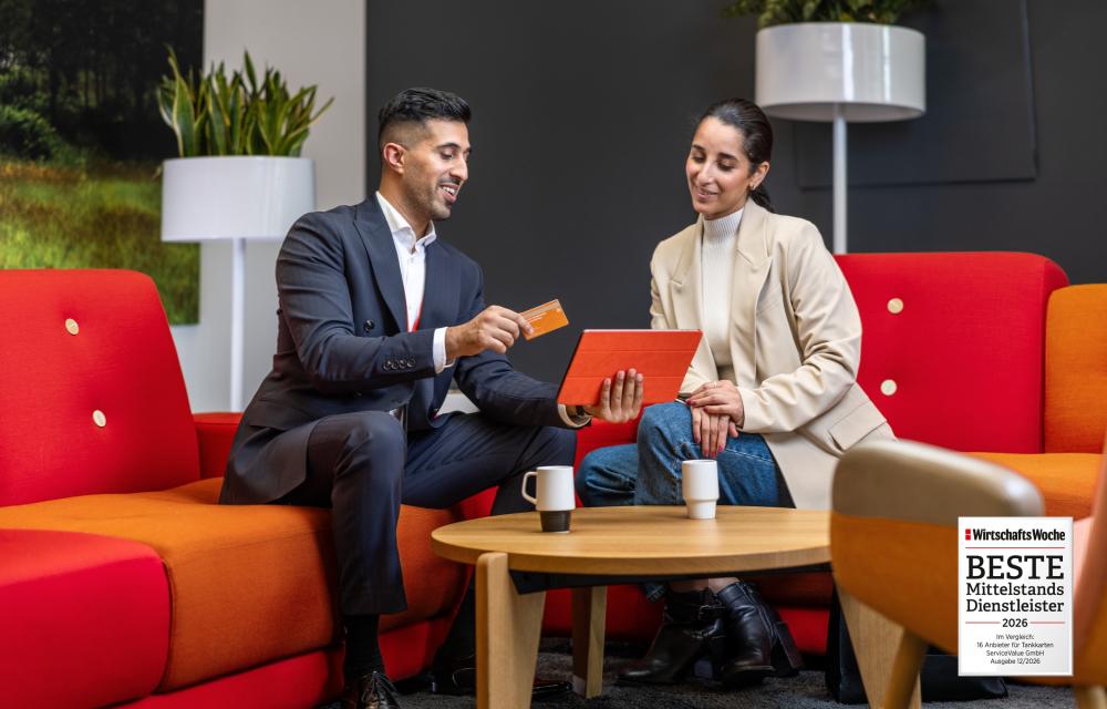Ein Mann im Anzug zeigt einer Frau im hellen Jacket die Vorteile der Tankkarte sitzend auf einem roten Sofa. Ein Siegel für den besten Mittelstandsdienstleiter 2026 ist unten rechts platziert.