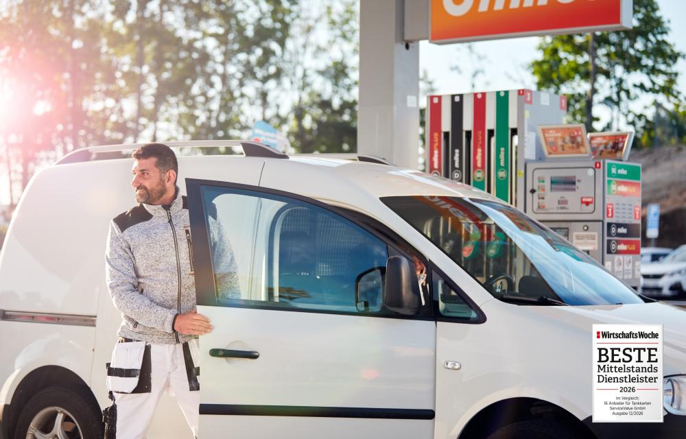 Ein Mann steigt aus einem weißen Kleinbus und steht vor einer Zapfsäule. Ein Siegel für den besten Mittelstandsdienstleiter 2026 ist unten rechts platziert